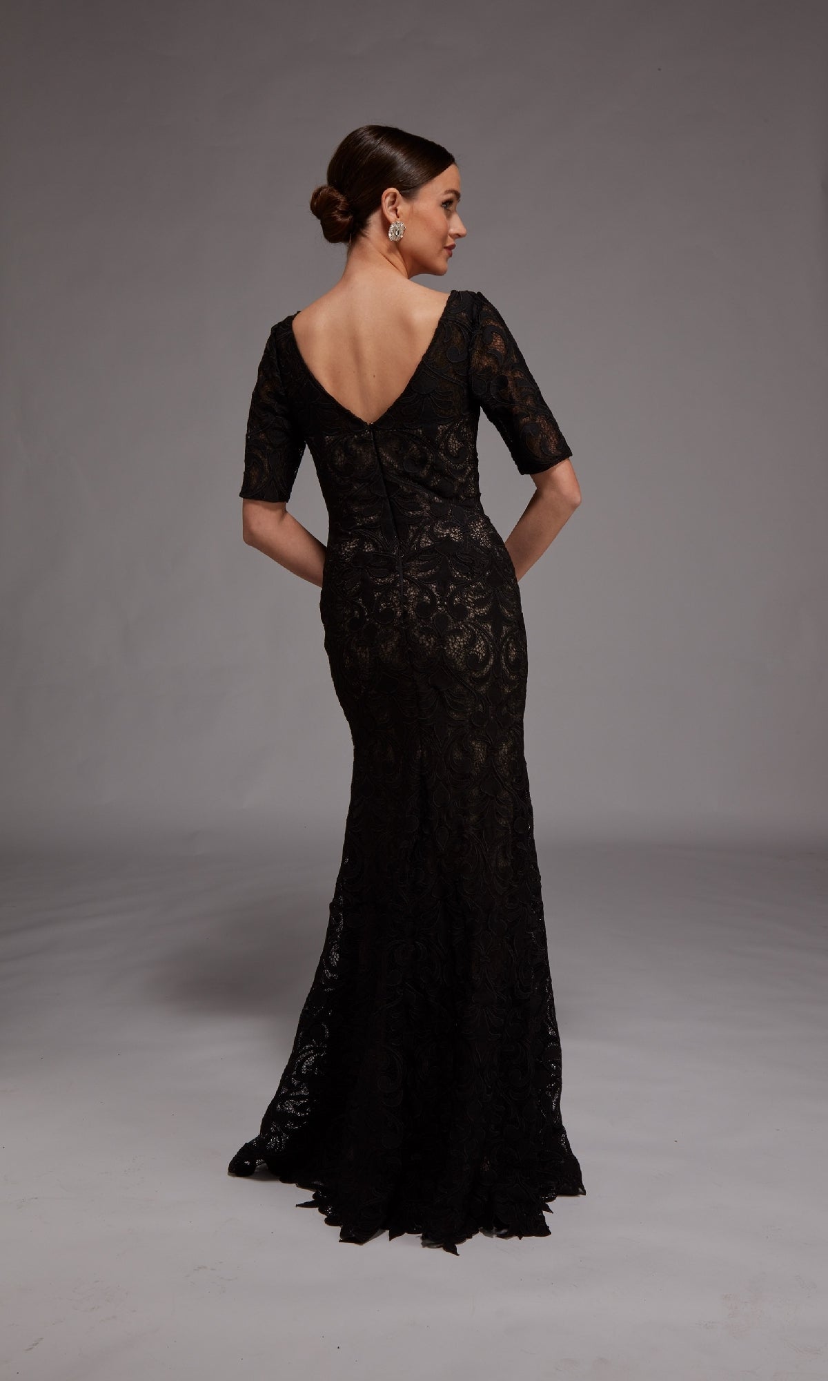 A woman faces away from the camera, wearing the Alyce 27871 long formal dress—a black lace overlay gown with short sleeves and a V-shaped back.